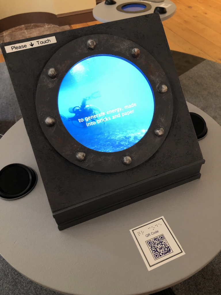An overhead 3/4 shot from above the Porthole station. A label that reads "Please Touch" is on the upper left corner of the porthole, and a QR code is on the pedistal. The Hot station is visible in the background. Photo credit Stefanie Koseff