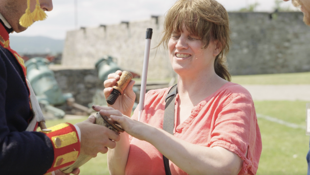 A woman with brown hair holding a long cane smiles as she touches a reproduction 18th-century powder horn being held by a man in a blue and red coat. A stone fort and mortars are in the background.