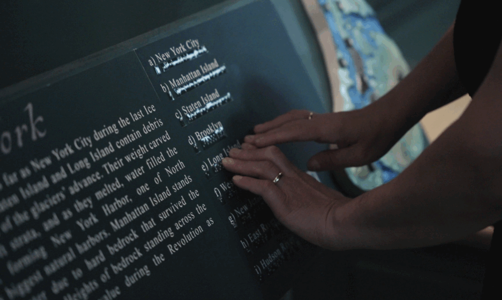 A close-up detail of two hands reading braille text "Long Island" on a label rail in an exhibit. The text identifies key features in a tactile map of New York that is visible in the background.